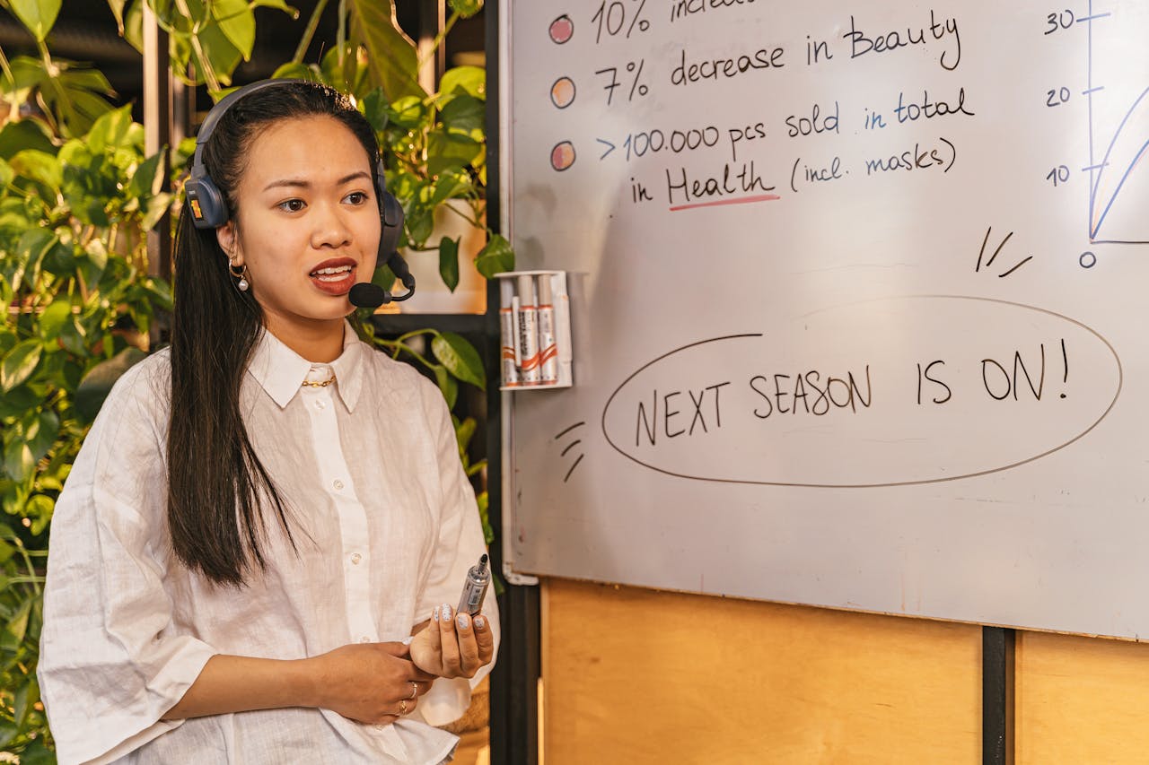 Woman presenting business statistics on whiteboard using headset, engaging audience.